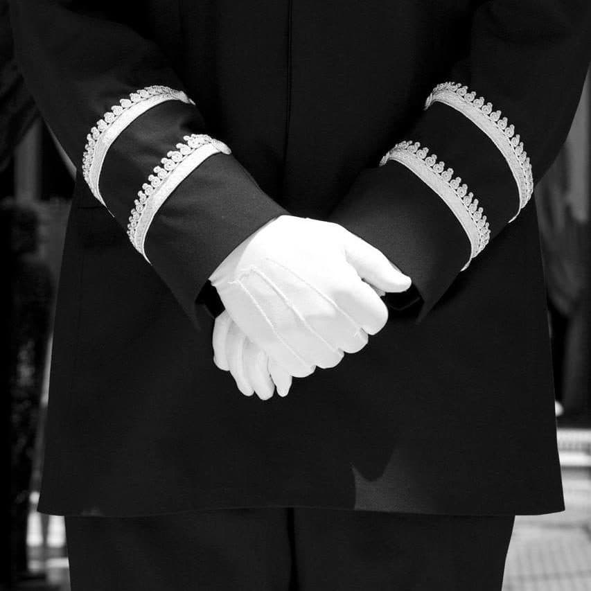 Butler in uniform, white gloves, hands crossed, black and white